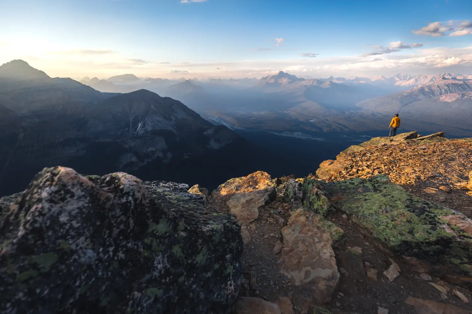 A person in a yellow jacket stands on the edge of Fairview Mountain, overlooking a vast mountain range under a clear sky. The mountains are bathed in warm, golden light from the setting sun, with rolling hills and distant peaks in the background.