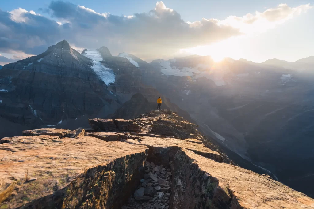 A person in a yellow jacket stands on Fairview Mountain, overlooking a dramatic landscape of snow-capped mountains under a cloudy sky with the sun setting in the distance.