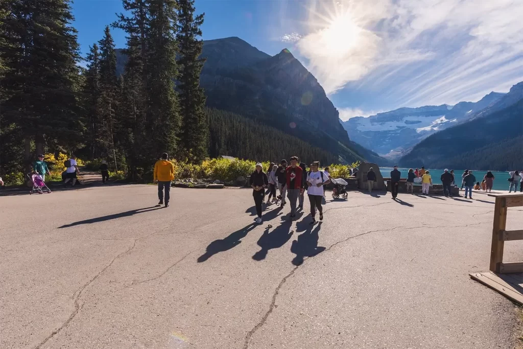 A group of people walk along a paved path with a mountain and forest backdrop. The sun shines brightly, casting long shadows. Lake Louise is visible in the distance, surrounded by trees and mountains. The sky is mostly clear with some clouds.