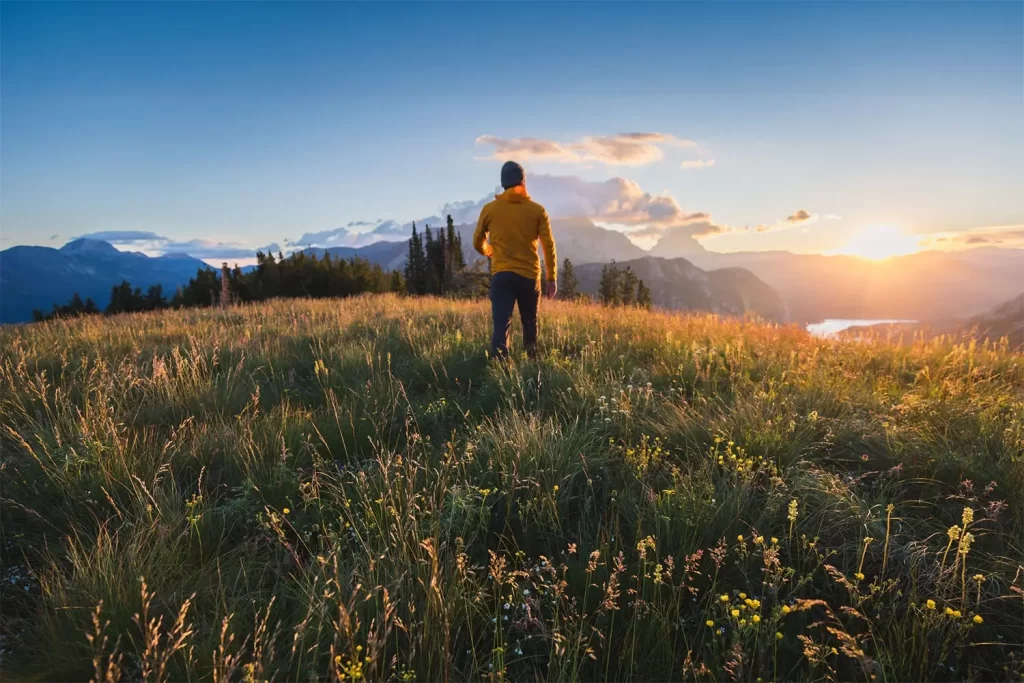 A person in a yellow jacket stands on a grassy hilltop, gazing at a scenic mountain landscape during sunset. The sky is partly cloudy, and the sun casts a warm glow over the scene.