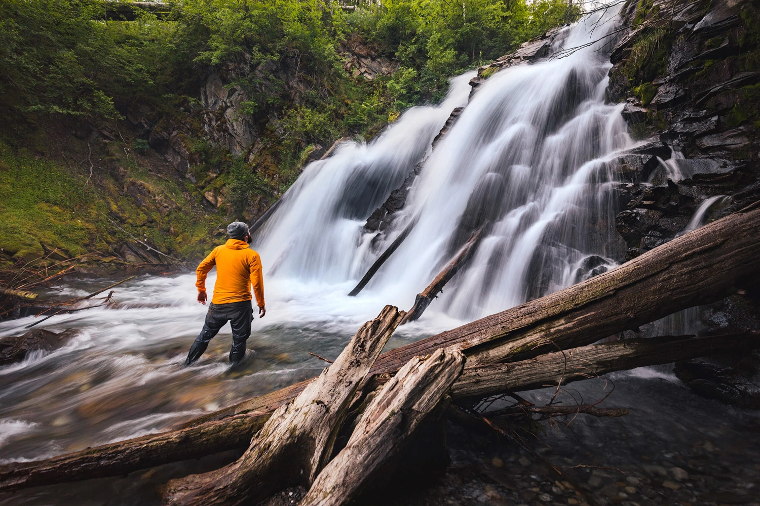 A person wearing a bright orange jacket stands near the base of Fairy Creek Falls, a wide, cascading waterfall surrounded by rocks, fallen logs, and green foliage.