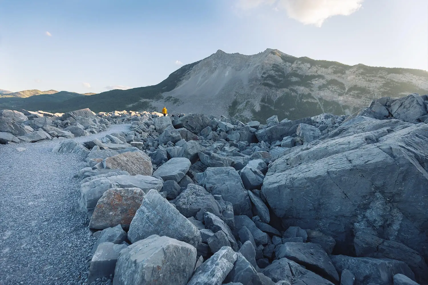 A rocky mountain landscape with large boulders and a gravel path in the foreground. A person in a yellow jacket stands on the distant Frank Slide Trail, with a tree-lined mountain rising under a clear sky.