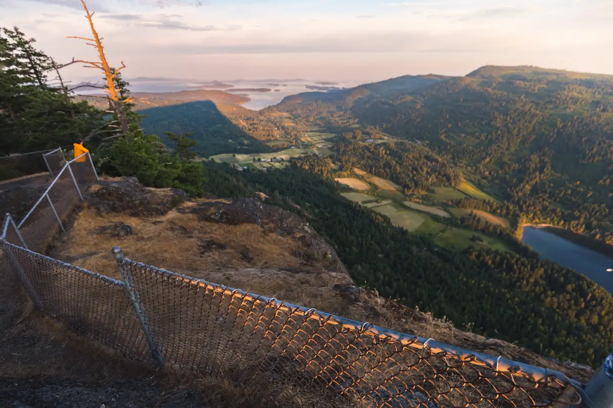 Experience the breathtaking view from a fenced lookout point on a mountain, overlooking fields, trees, and distant water beneath a sunset sky—one of the most memorable Salt Spring Island hikes.