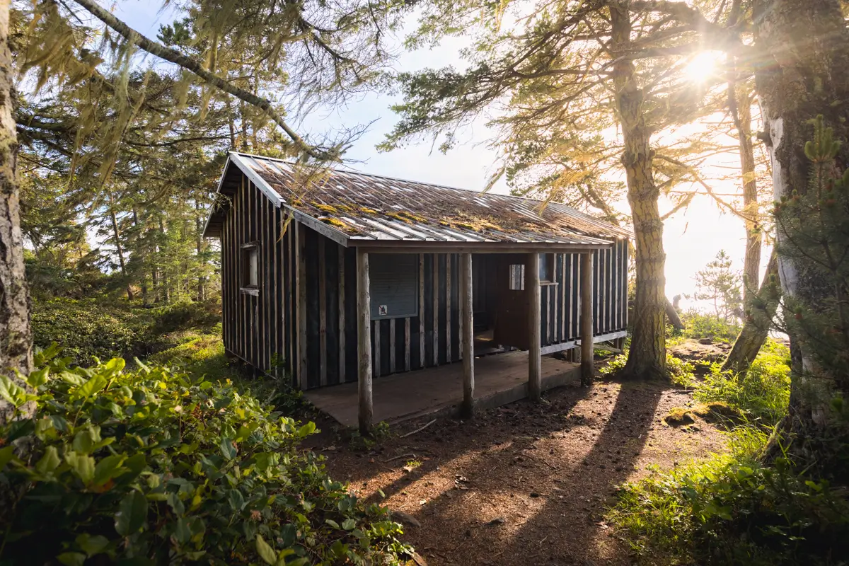 A small wooden cabin with a covered porch sits nestled among trees and greenery near the East Sooke Coast Trail. Sunlight filters through the branches, casting shadows on the ground and illuminating parts of the cabin and forest.