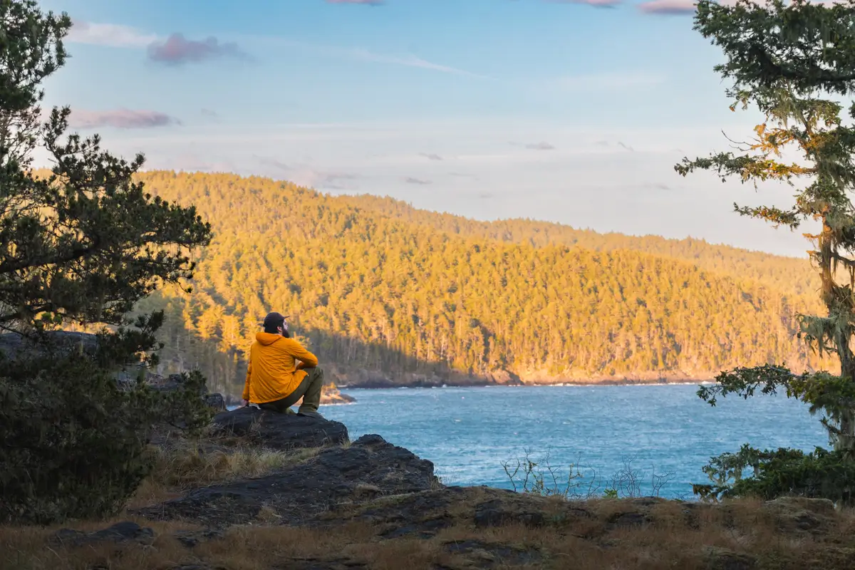 A person in a yellow jacket sits on a rocky outcrop overlooking the water along the East Sooke Coast Trail, with forested hills in the background under a partly cloudy sky. Trees frame the scene on either side.