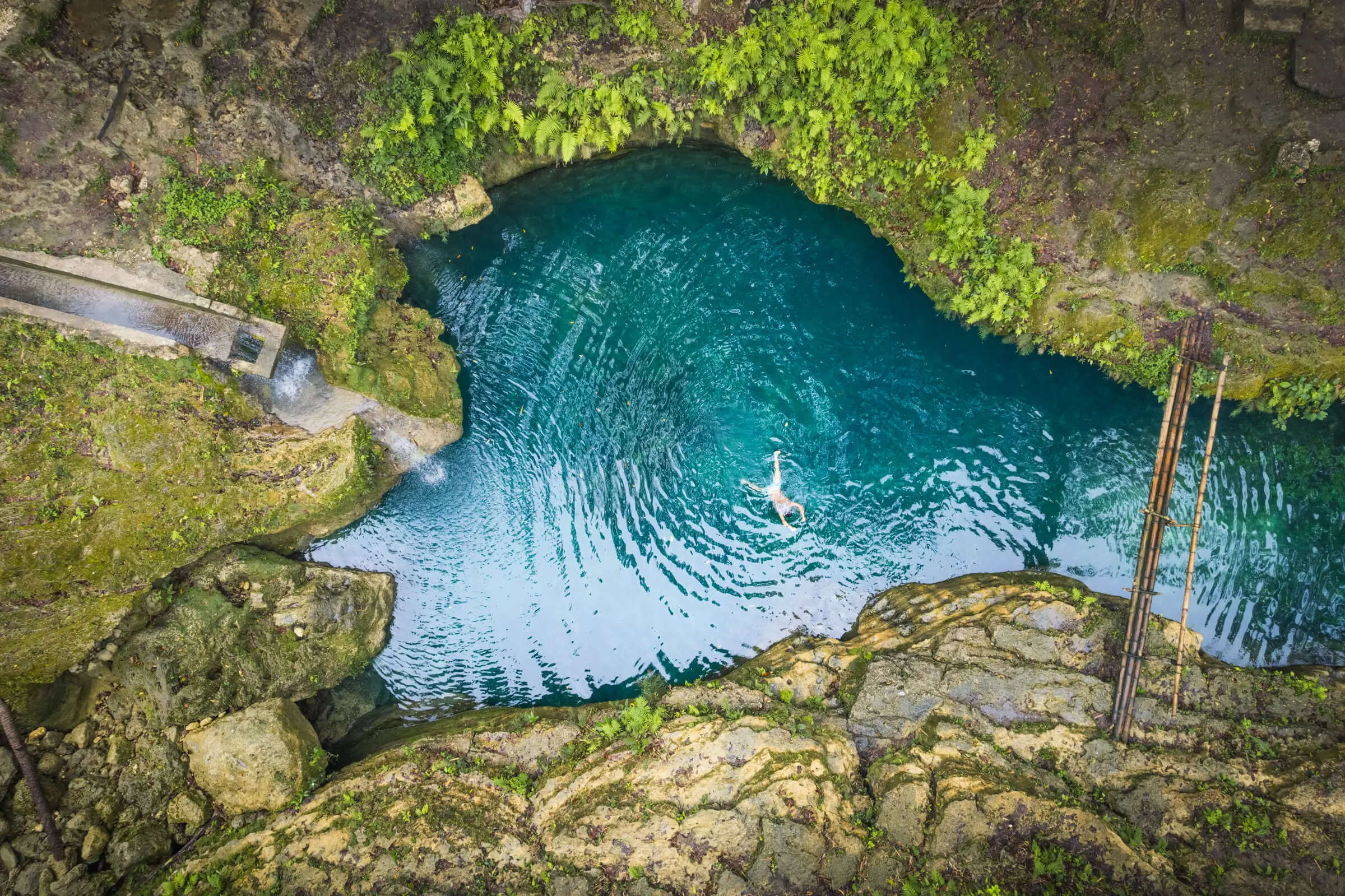 A person swims in a small, clear blue natural pool surrounded by rocky terrain and green vegetation, viewed from above—one of the hidden gems among Moalboal tourist spots.