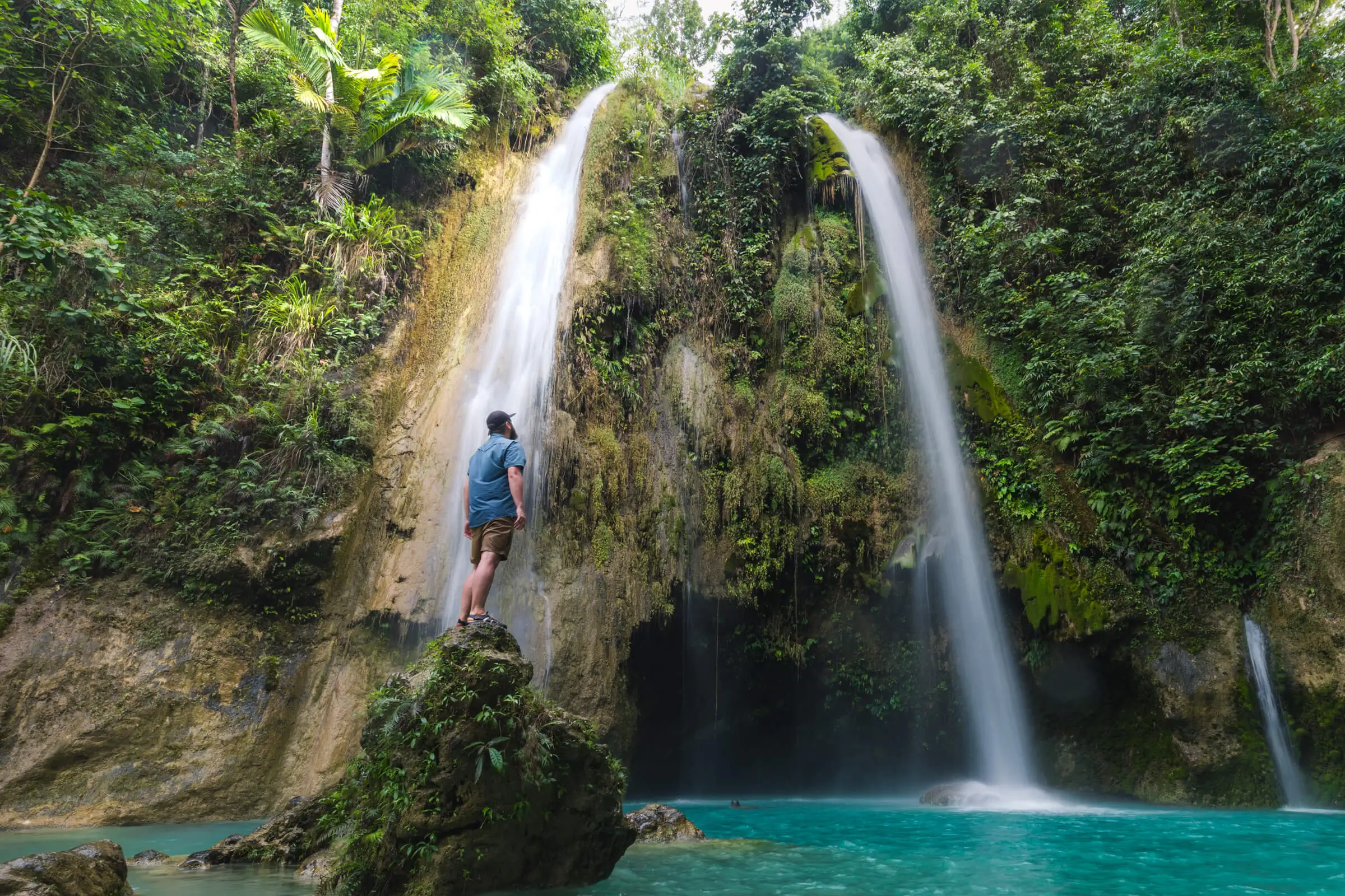 A person stands on a rock in a turquoise pool, facing two tall waterfalls flowing down a mossy cliff surrounded by dense green tropical vegetation—a breathtaking scene among Moalboal tourist spots.