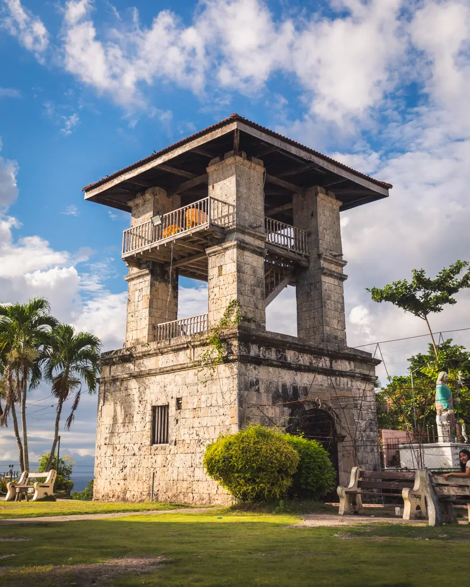 A stone watchtower with a tiled roof and balconies stands on a grassy area with benches and palm trees, making it one of the charming Moalboal tourist spots against a partly cloudy sky.