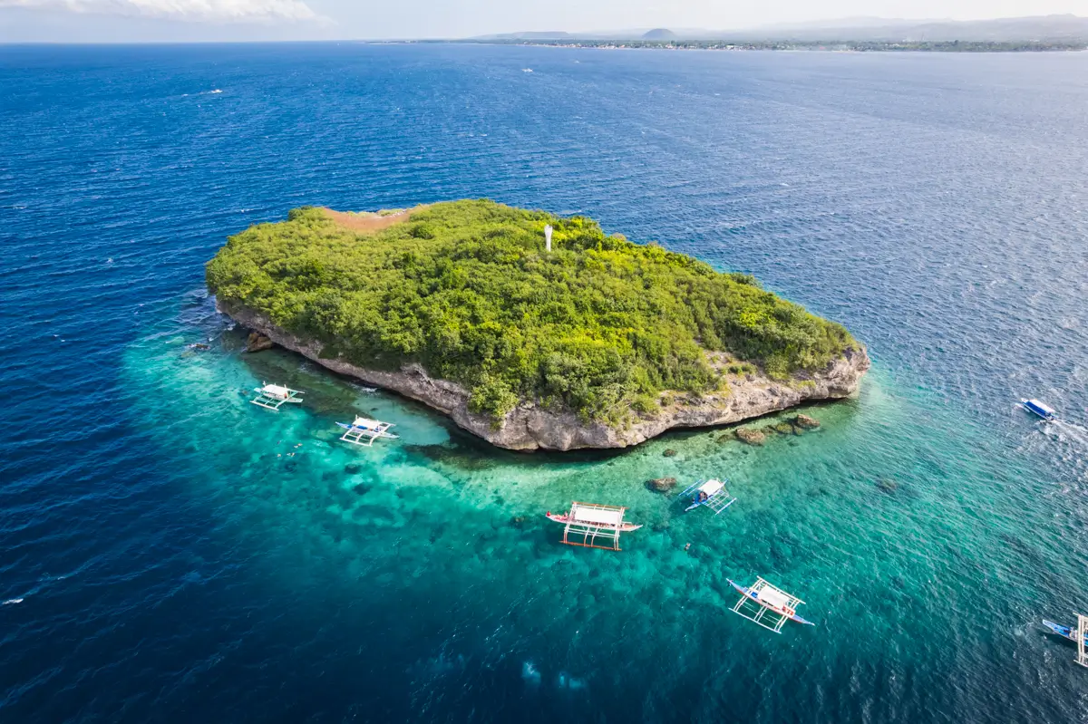 An aerial view of a small, lush green island surrounded by clear blue water, with several white boats anchored near the shore—one of the picturesque Moalboal tourist spots. The island features rocky edges and a single structure near its center.