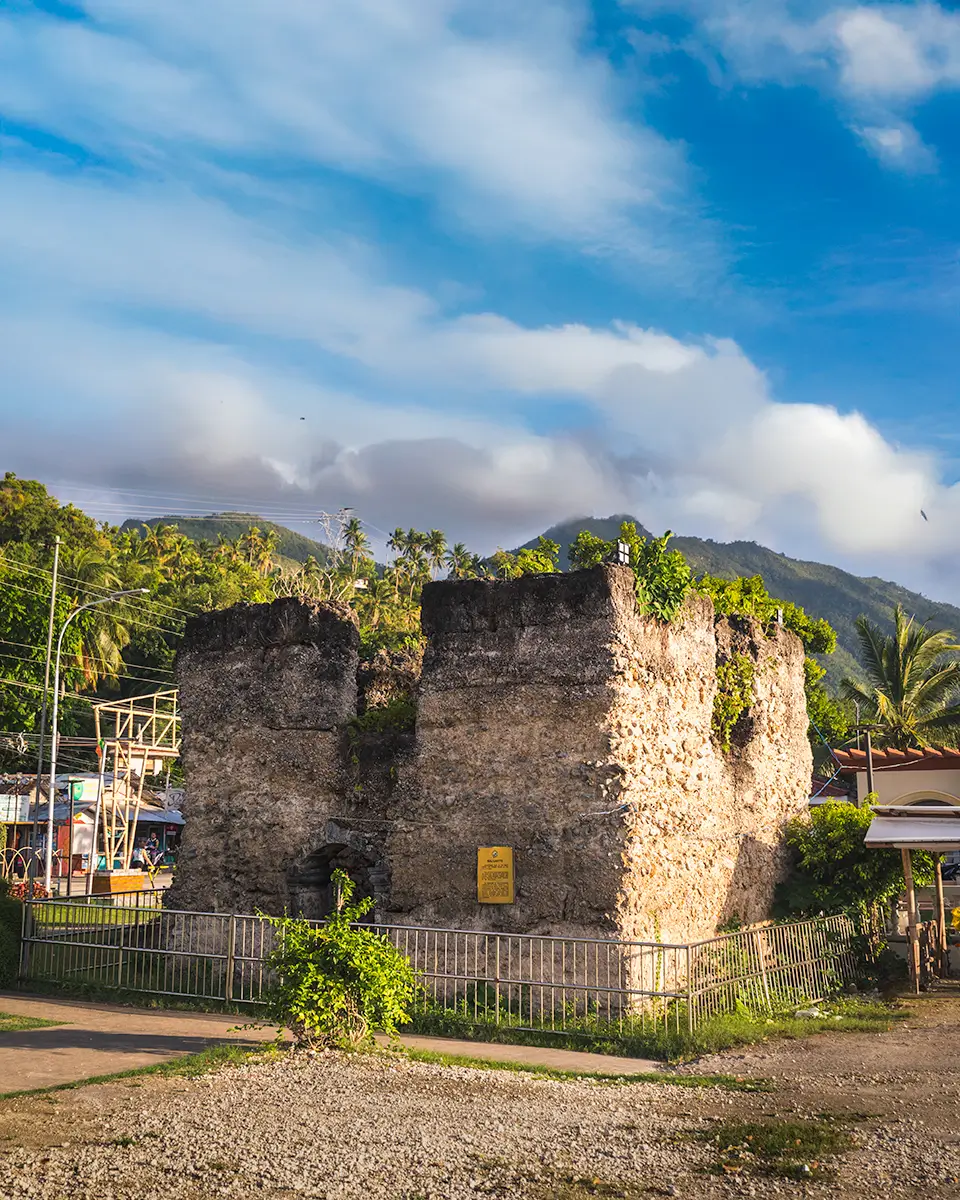 A small, square stone fort with weathered walls is surrounded by a metal fence. This historic site, one of the notable Moalboal tourist spots, features a front wall sign and is set against palm trees, buildings, and distant mountains under a partly cloudy sky.