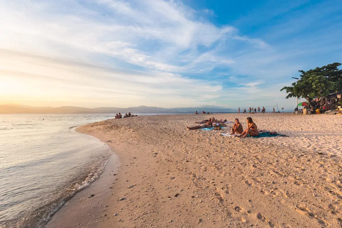 People relax on blankets and towels on a sandy beach at sunset—one of the serene Moalboal tourist spots. Small groups are spread along the shore, with a few people standing near the calm sea as clouds drift across a clear sky.