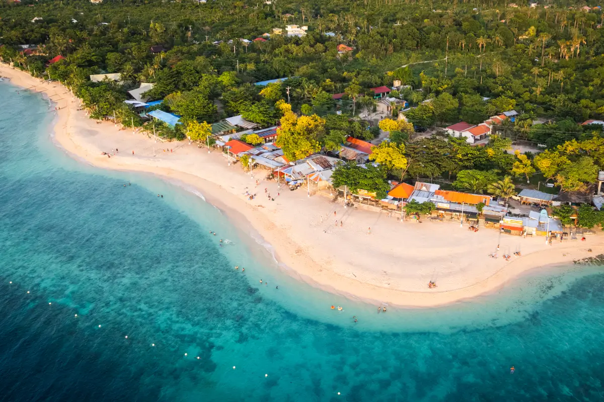 Aerial view of a curved sandy beach lined with small buildings and trees, featuring clear blue and turquoise water. People enjoy the shoreline, one of the stunning Moalboal tourist spots surrounded by lush green vegetation.
