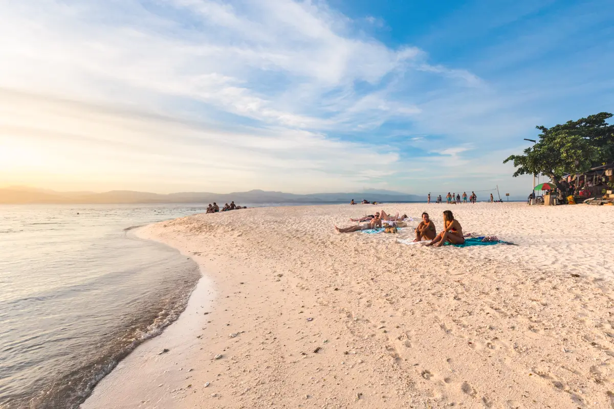 People relax on a sandy beach near the shoreline during sunset, with calm water and a group gathered in the background. Trees and small structures line the right—a perfect scene among Moalboal tourist spots.