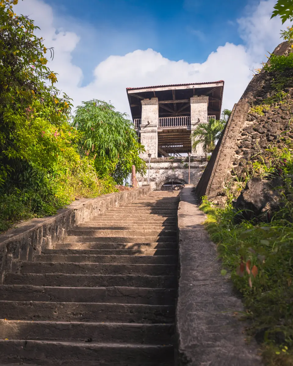Stone steps lead up to a two-story stone structure with a metal roof, surrounded by green foliage and set against a blue sky—an inviting scene reminiscent of Moalboal tourist spots.