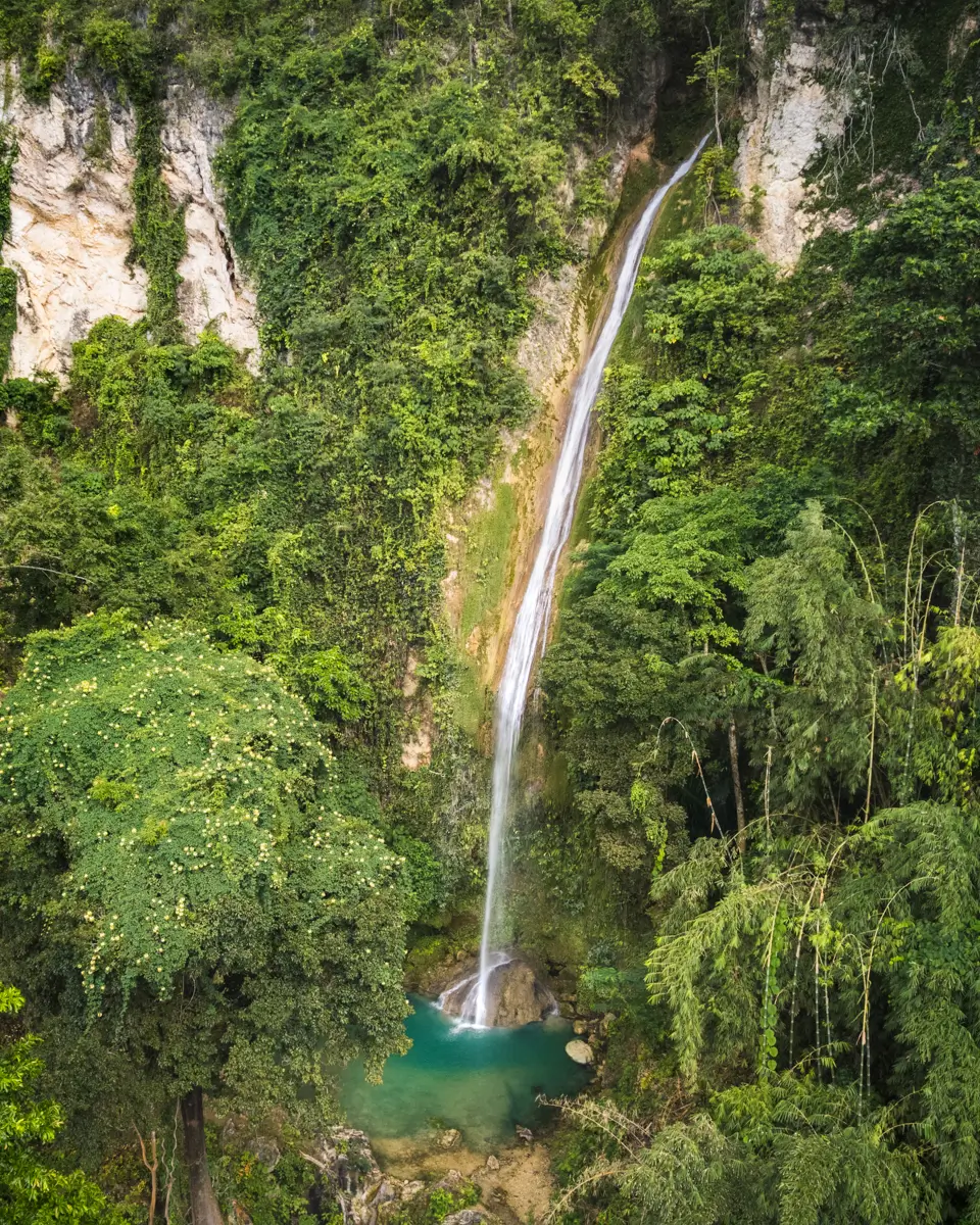 A tall, narrow waterfall cascades down a steep, rocky cliff covered in dense green vegetation—one of the stunning Moalboal tourist spots—ending in a small turquoise pool surrounded by lush trees and plants.