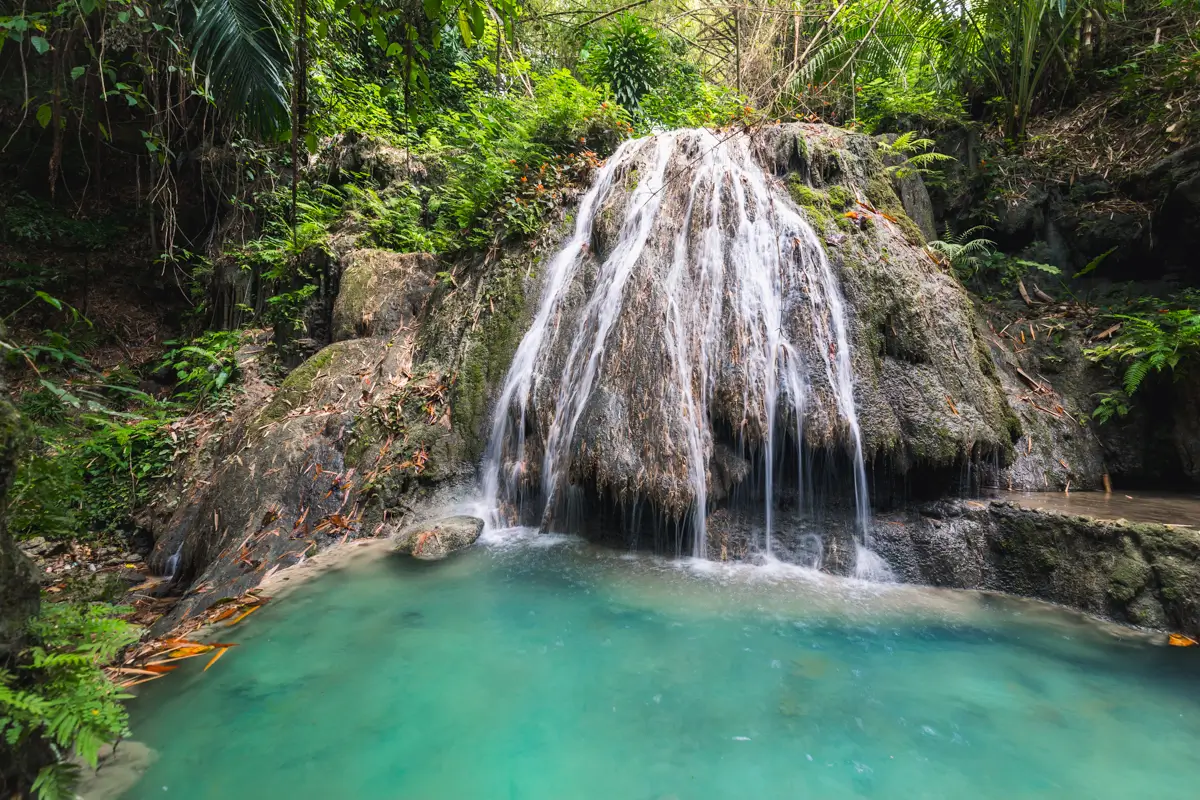 A small waterfall flows over mossy rocks into a clear turquoise pool, surrounded by dense green vegetation—a hidden gem among Moalboal tourist spots in this lush tropical forest setting.