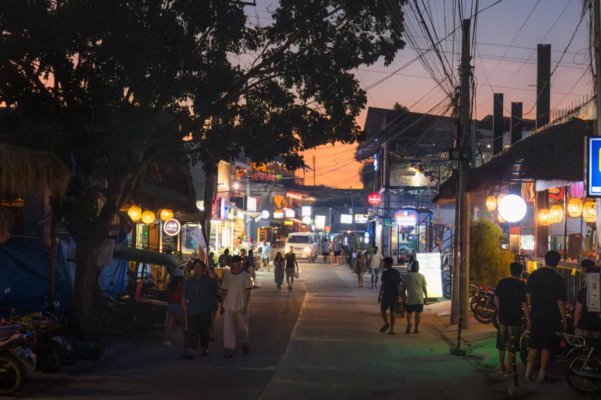 A lively street scene at dusk in Moalboal tourist spots shows people walking along a road lined with shops, restaurants, and bright signs. Trees, power lines, and bicycles are visible, with buildings illuminated by various lights.