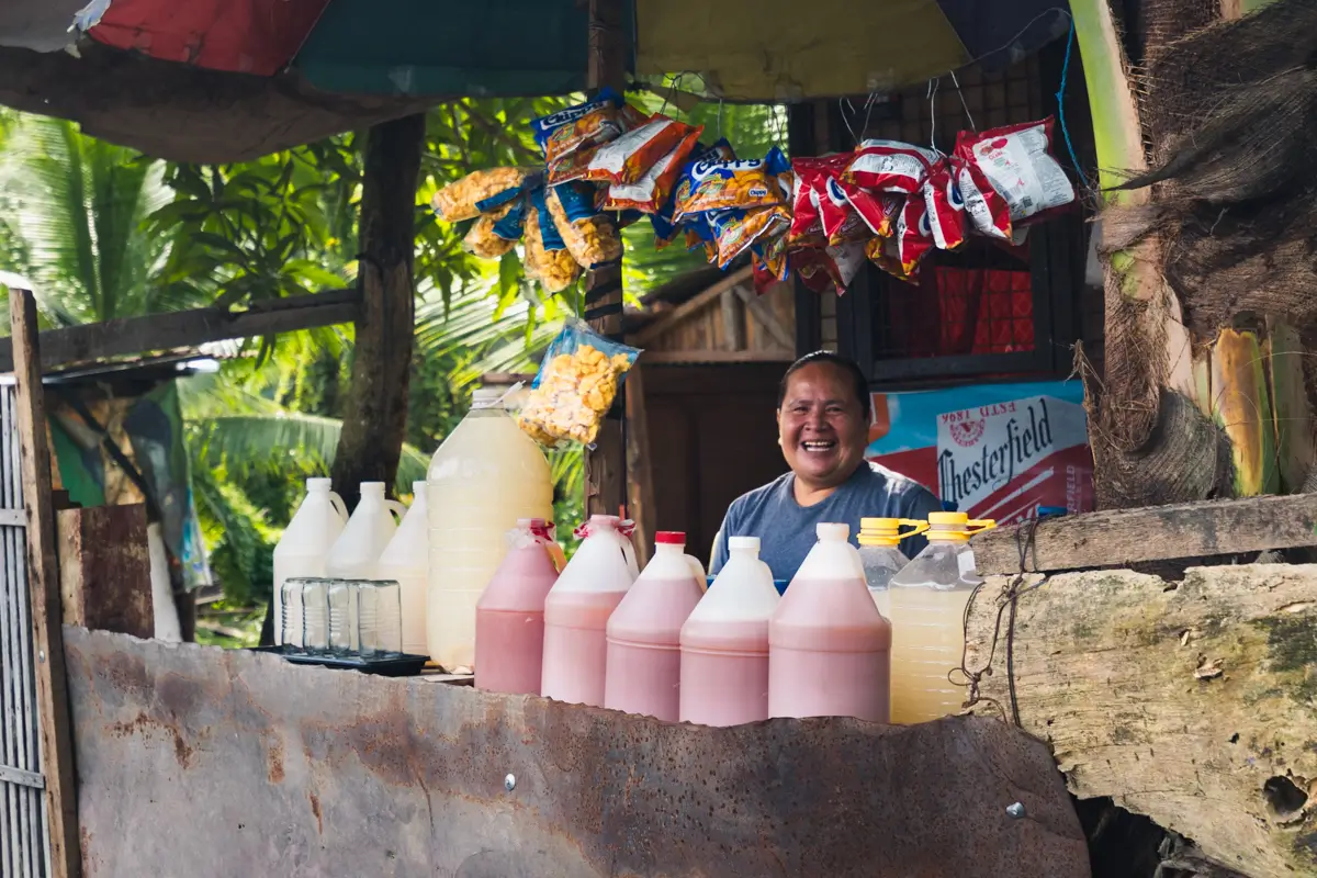 A person stands at a rustic roadside stall near popular Moalboal tourist spots, surrounded by large plastic bottles of pink and white drinks, hanging snack packs, and lush greenery in the background.
