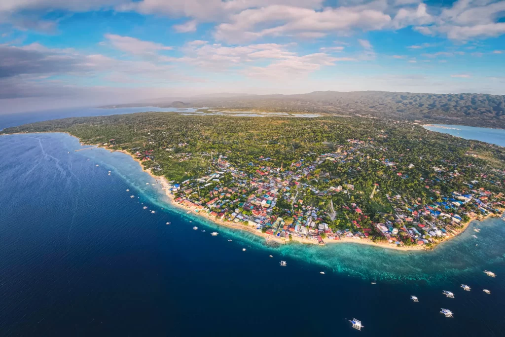 Aerial view of a coastal town, featuring dense greenery and buildings near the shoreline, bordered by deep blue ocean waters dotted with small boats—showcasing Moalboal tourist spots under a partly cloudy sky.