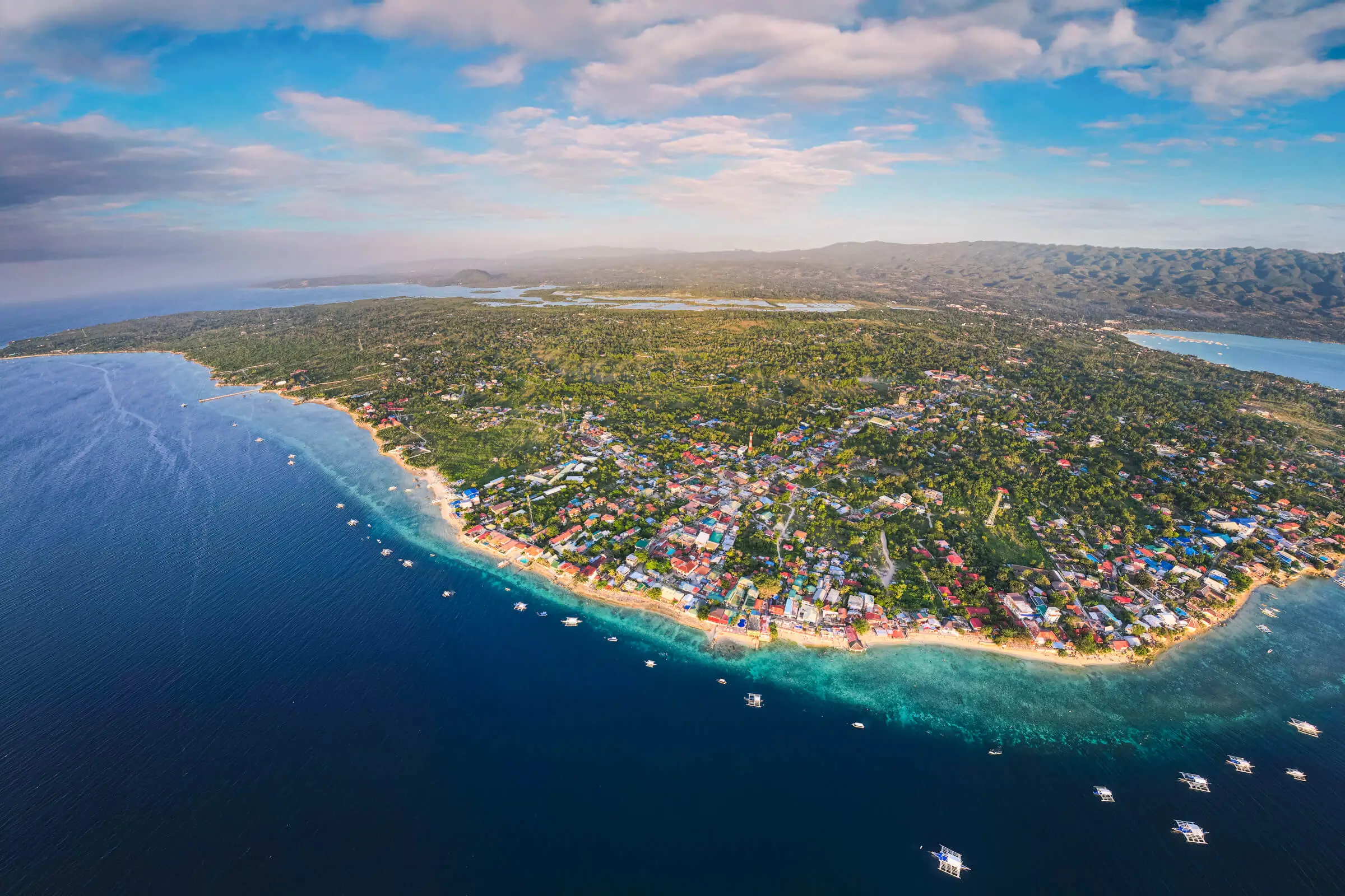Aerial view of a coastal town, featuring dense greenery and buildings near the shoreline, bordered by deep blue ocean waters dotted with small boats—showcasing Moalboal tourist spots under a partly cloudy sky.