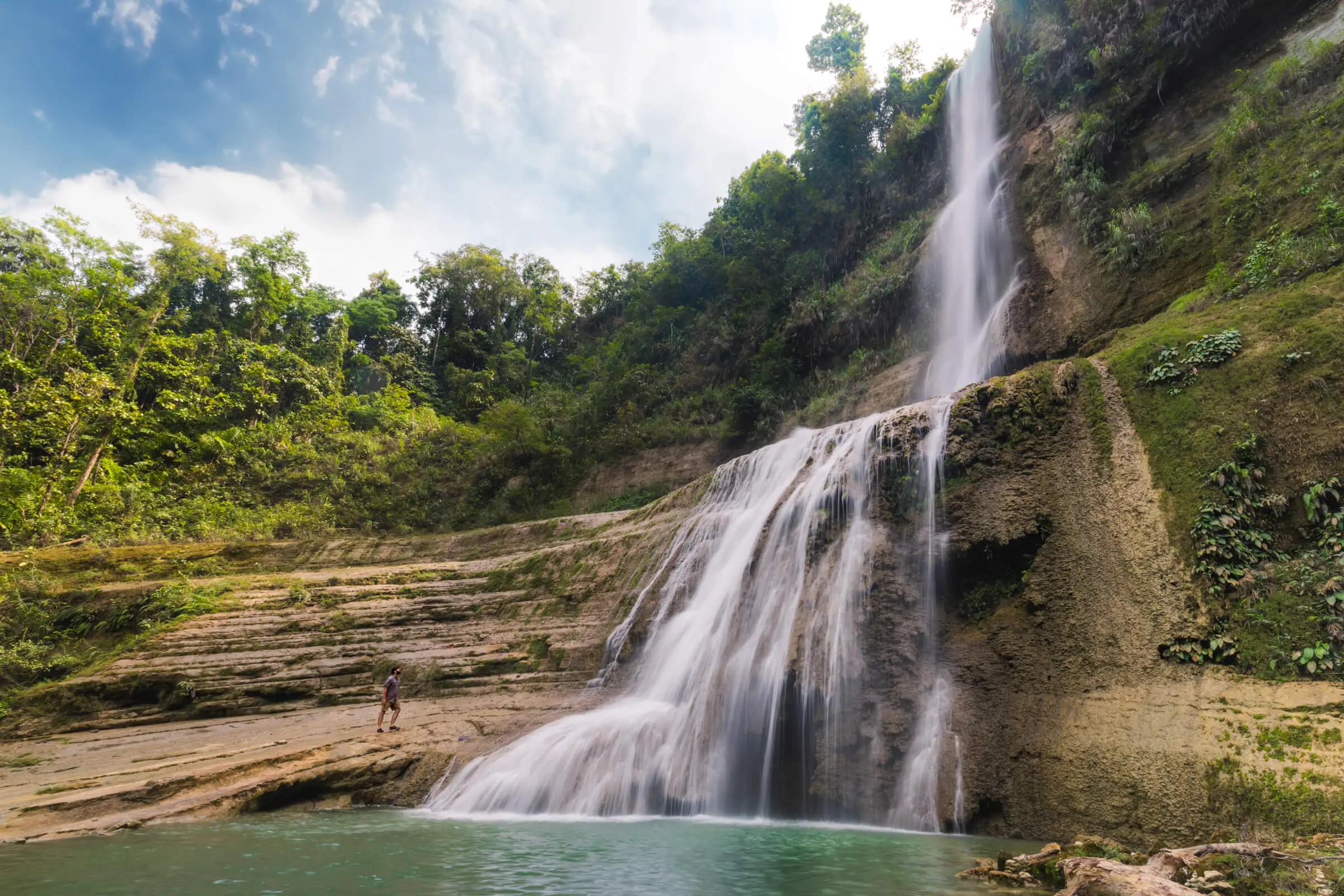 A tall waterfall, known as Pahangog Falls, cascades down a rocky cliff surrounded by lush green vegetation, with a person standing near the water’s edge for scale. The water flows into a turquoise pool at the bottom.