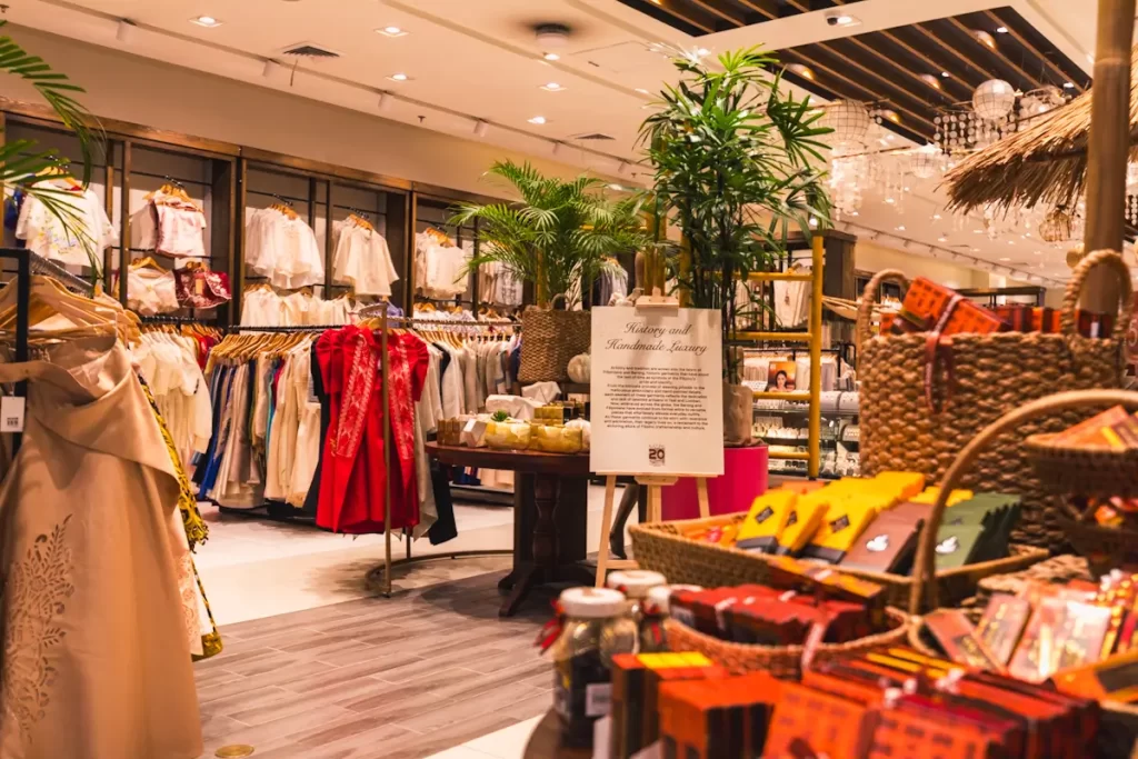 A retail store interior displays clothing on racks, baskets of packaged goods, and decorative elements like potted plants under bright lighting, creating a warm atmosphere reminiscent of Cebu City tourist spots.