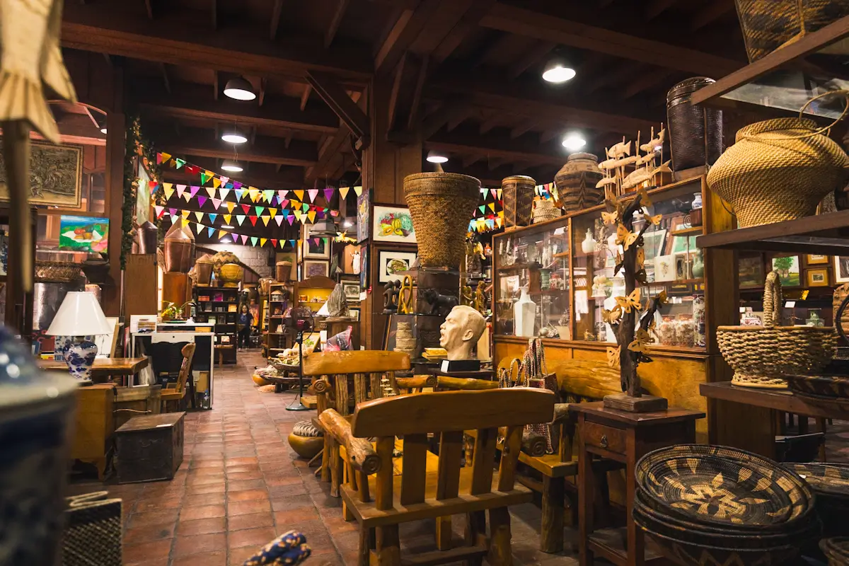 A cluttered antique shop interior with wooden furniture, woven baskets, and shelves filled with vintage items. Colorful triangular flags hang overhead, and warm lighting highlights this hidden gem among Manila tourist spots.
