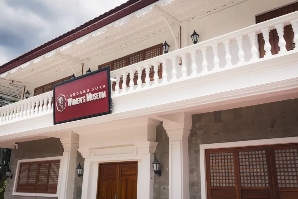 A two-story building with white railings and columns features a red sign that reads Tandang Sora Womens Museum above the entrance—one of the notable Manila tourist spots. Wooden grids accent the doors and windows, with wall-mounted lamps flanking the doorway.