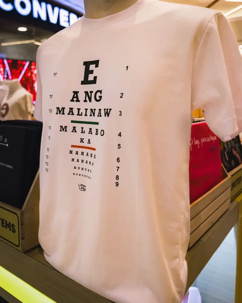 A white T-shirt displayed on a mannequin features a printed eye chart design with the words E ANG MALINAW MALABO KA in decreasing font size, perfect for those exploring Manila tourist spots with a touch of humor.