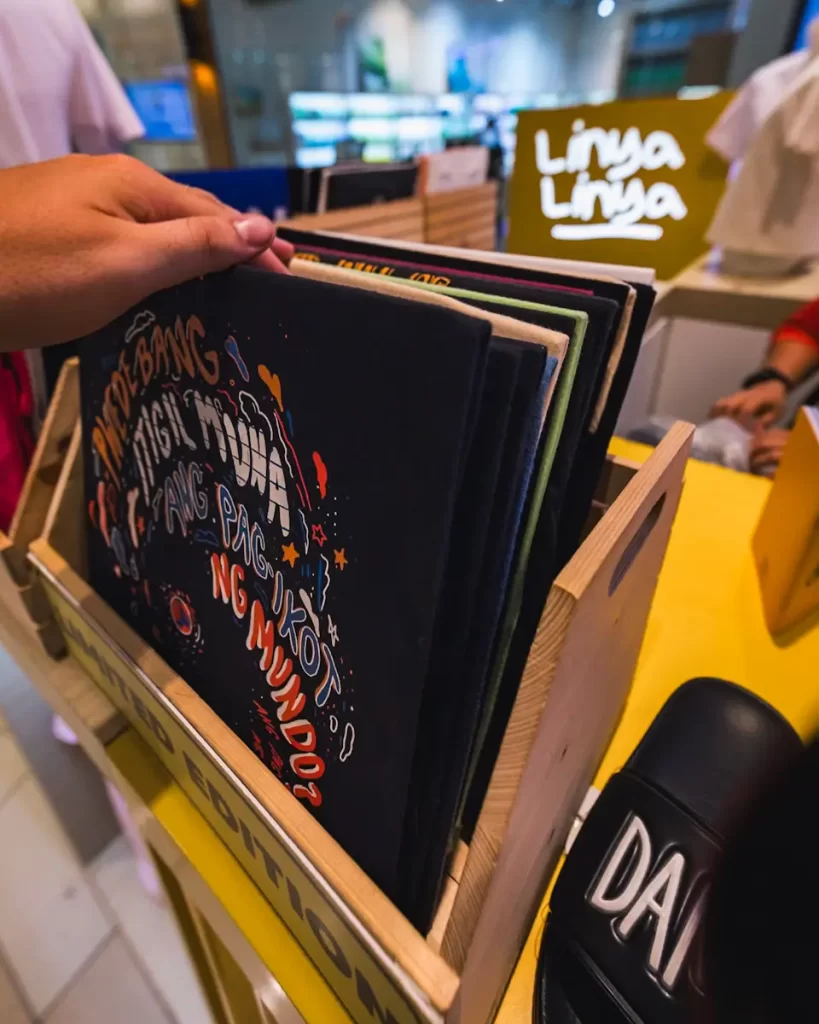 A person flips through a wooden crate of graphic T-shirts at a store display, reminiscent of the creative shops found near Manila tourist spots. A yellow sign in the background reads “Linya Linya,” with shirts and fixtures surrounding the area.