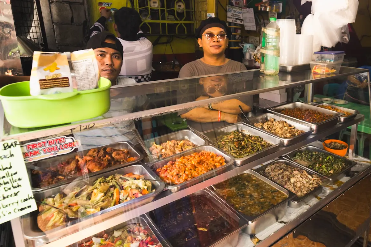 Two people stand behind a glass food counter displaying various prepared dishes in a casual, brightly lit eatery—an inviting stop for those exploring Manila tourist spots, with kitchen supplies and handwritten signs in the background.