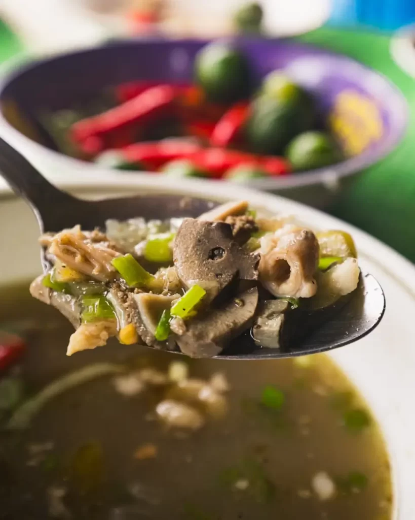 A close-up of a spoonful of soup with chopped meat, green onions, and vegetables above a bowl of broth—reminiscent of flavors found near popular Manila tourist spots. In the background, whole red chilies and small green limes add color.