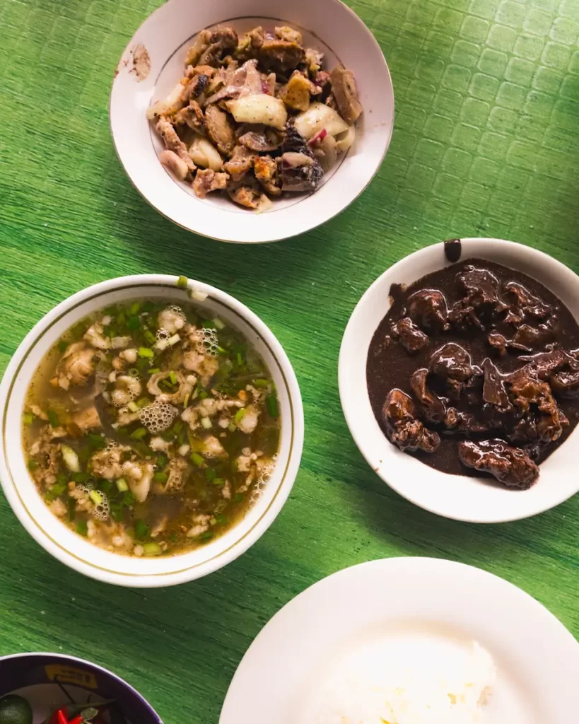 Three dishes are placed on a green textured table: a bowl of soup with meat and herbs, a plate of dark brown sauced meat, and a plate of cooked chicken pieces—flavors often found near famous Manila tourist spots. There is also a partial view of white rice.
