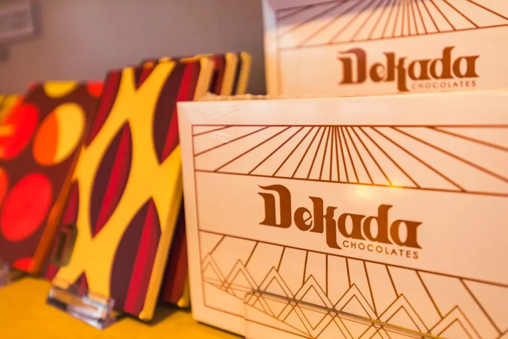 Two boxes of Dekada Chocolates are on display next to several colorful chocolate bars with geometric patterns in red, yellow, and purple tones—perfect sweet souvenirs from your visit to Manila tourist spots. The boxes have brown and gold decorative designs.