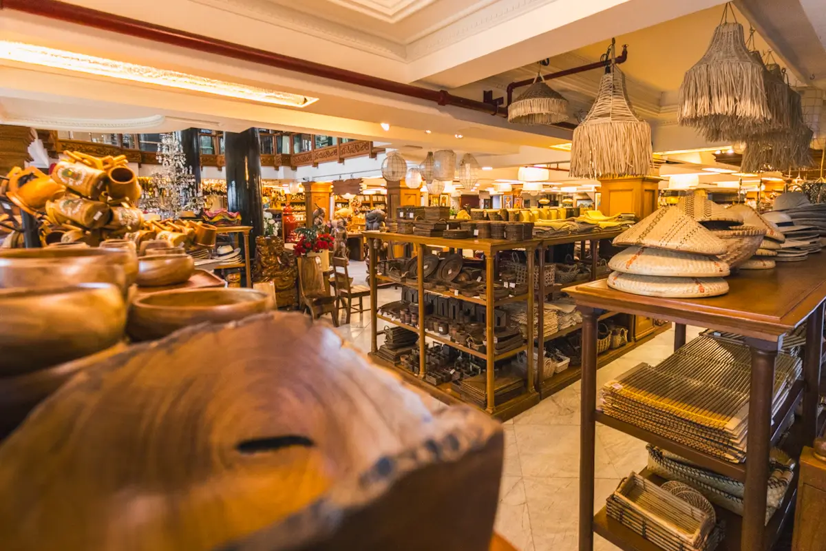 A store interior displays various wooden bowls, woven baskets, and handicrafts on shelves and tables—perfect souvenirs from Manila tourist spots. Pendant lights hang from the ceiling above the tiled floor in this well-lit, organized shop.