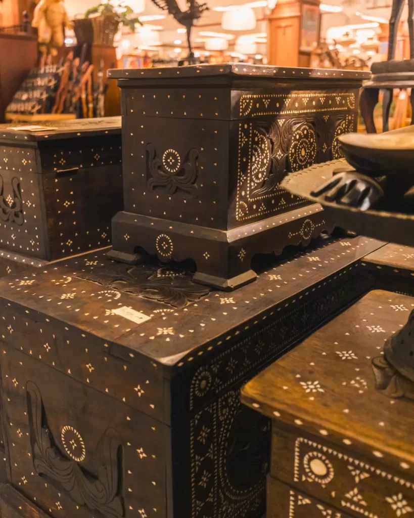 Wooden chests with intricate white inlay patterns are stacked in a store, surrounded by other furniture and decorative items in a warmly lit indoor setting—capturing the charm often found in Manila tourist spots.
