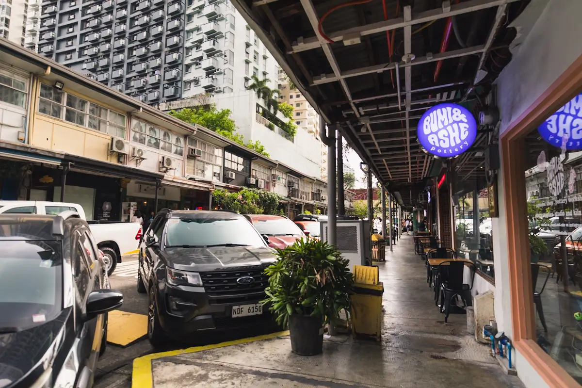 A city street scene with parked cars, potted plants, and a covered sidewalk in front of stores. A purple neon sign reads “Burn & Ash.” High-rise and low-rise buildings frame the view, capturing the vibe of Manila tourist spots.