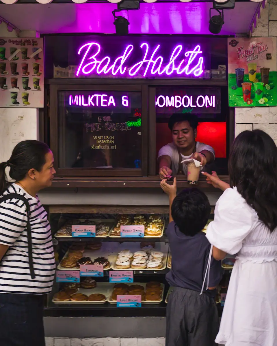 A man hands a drink to a child at a small kiosk named Bad Habits with a neon sign. This cozy spot, reminiscent of hidden Manila tourist spots, has three people at the counter and tempting pastries displayed in glass cases below.