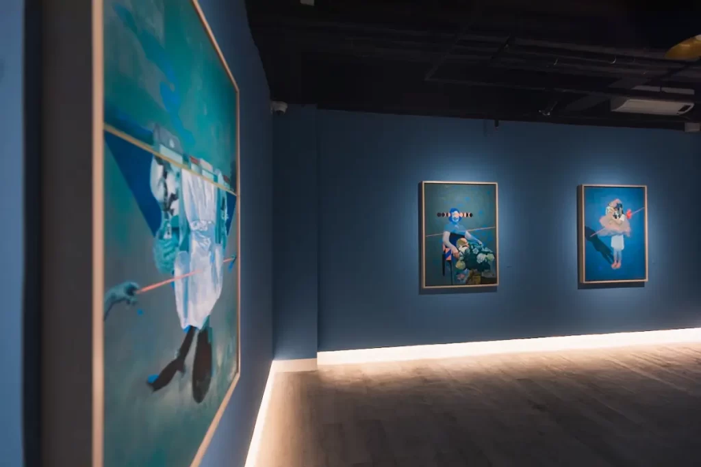A room with blue walls displays three framed paintings, each featuring abstract human figures with blue tones. The gallery, reminiscent of Manila tourist spots, has wood flooring and minimal lighting along the base of the walls.