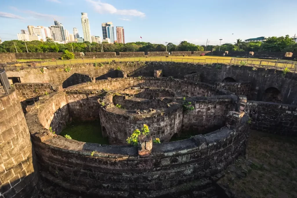 A stone circular fortification with overgrown vegetation, part of an old walled structure, stands in the foreground—a reminder of Manila tourist spots—while modern city buildings and open green space stretch into the clear sky beyond.