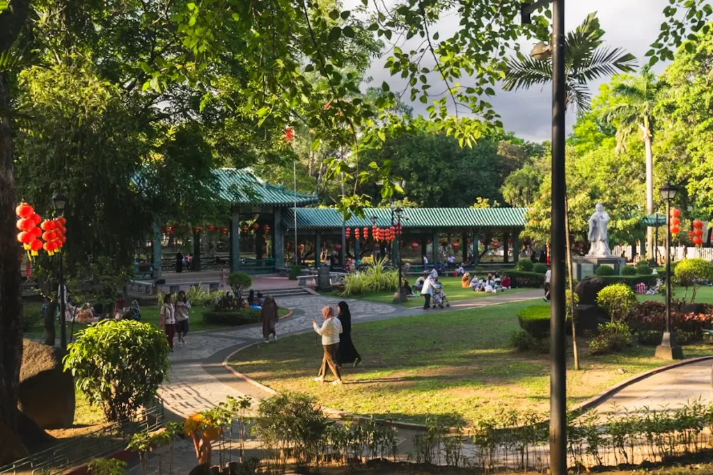People walk and relax in a lush, green park with trees, a covered pavilion, red lantern decorations, and a statue—one of the charming Manila tourist spots. Sunlight filters through the trees, casting shadows on the grass and pathways.