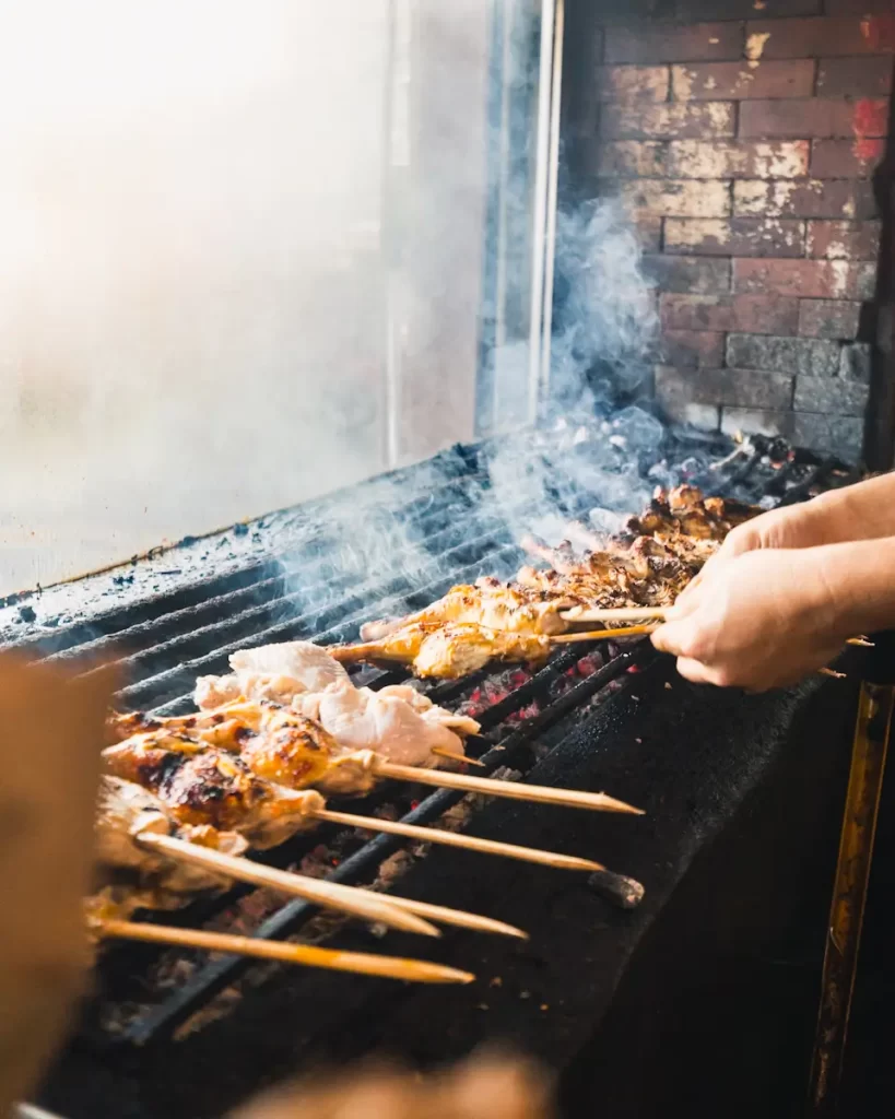 A person grills skewers of meat over an open flame on a charcoal grill, with smoke rising and a brick wall in the background—a scene reminiscent of street food found near Manila tourist spots. The skewers are at different stages of cooking.