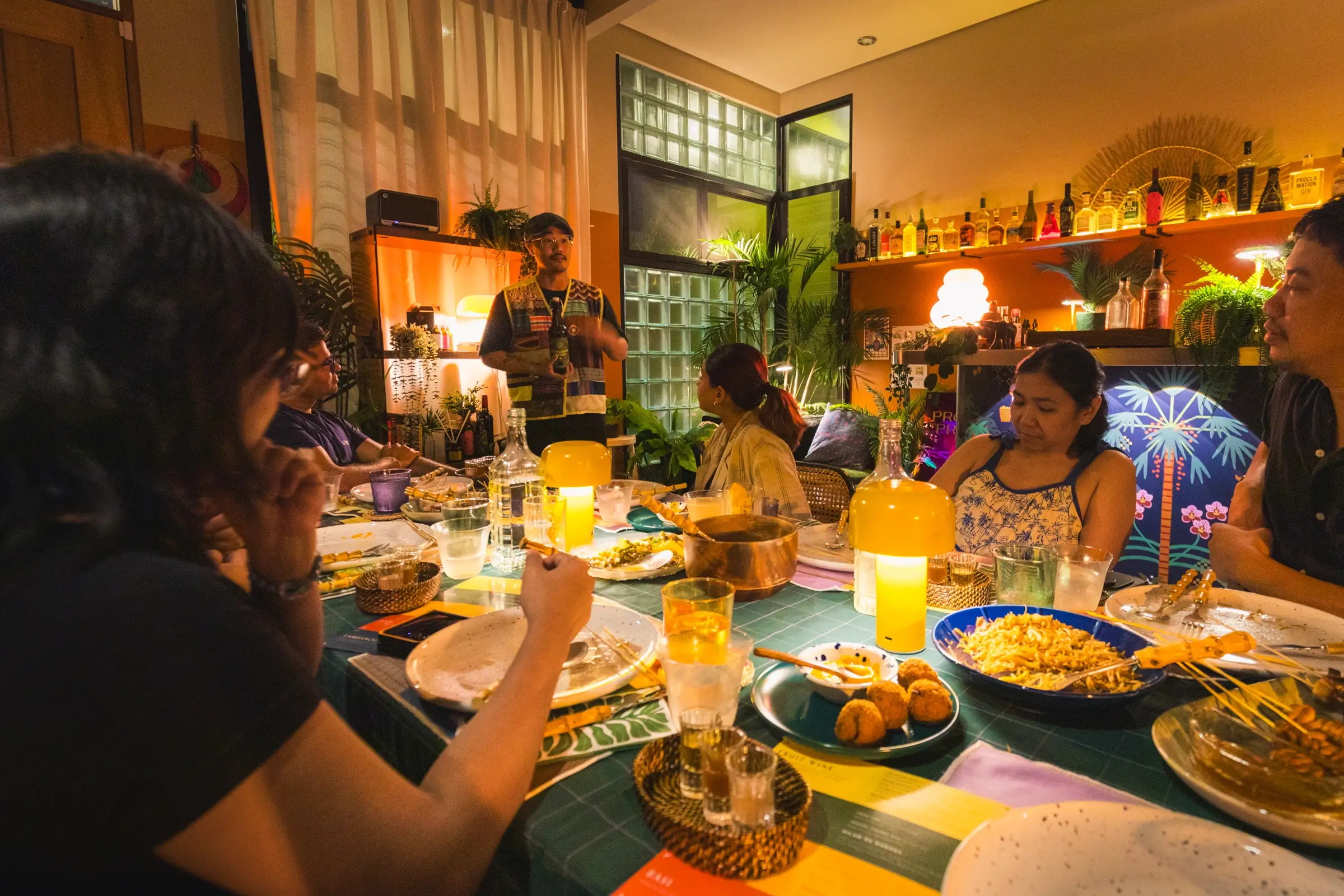 Six people are seated around a dining table set with various dishes and drinks, reminiscent of gatherings after exploring Manila tourist spots. One person stands speaking, while the warmly lit room is decorated with plants, bottles, and colorful decor.