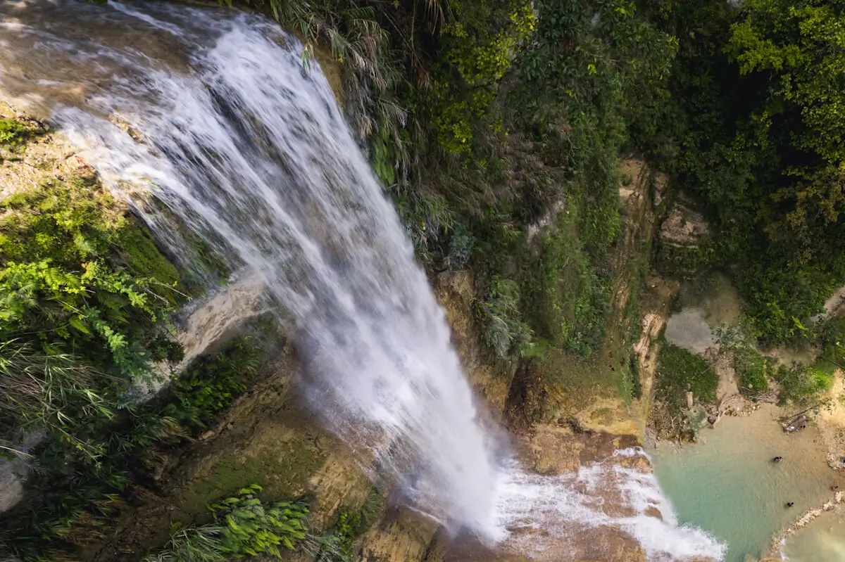 A waterfall flows over a rocky ledge into a pool below, surrounded by green vegetation and steep cliffs—an aerial view of the stunning Can-Umantad Falls.