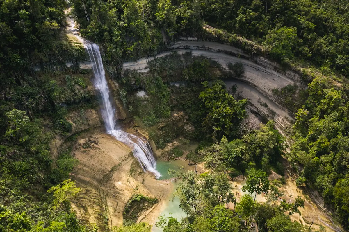 Aerial view of Can-Umantad Falls cascading down a rocky cliff surrounded by dense green forest, with water collecting in turquoise pools at the bottom.