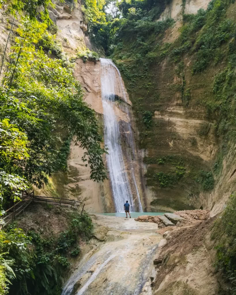 A person stands at the base of Dao Falls, surrounded by steep, rocky cliffs and dense green vegetation under daylight. A wooden railing lines the left side of the area near the waterfall.
