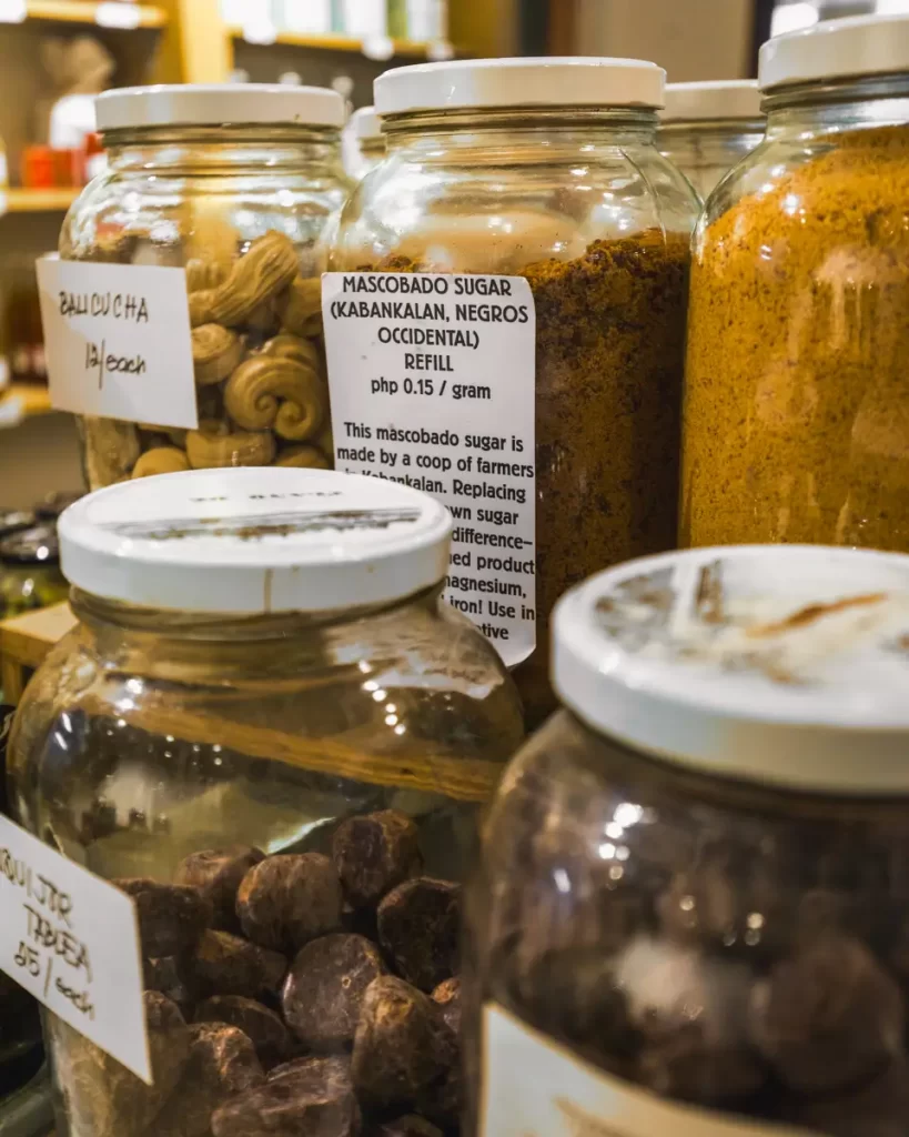 Several glass jars filled with various dried goods, including nuts and sugar, are displayed on a shelf—often found in shops near Dumaguete Tourist Spots. Each jar has a white label with handwritten or printed information about its contents and price.