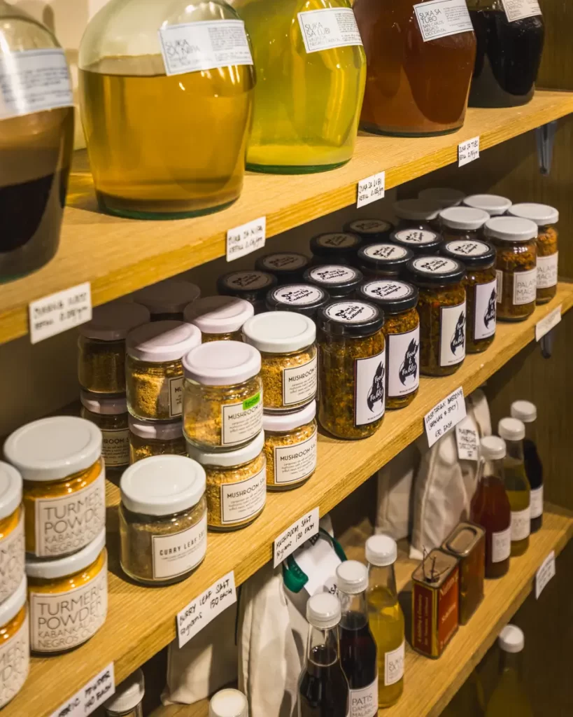 Wooden shelves display jars and bottles filled with various oils, powders, spreads, and sauces, each labeled with white tags. Reminiscent of finds at local Dumaguete tourist spots, the items are neatly organized and grouped by type.