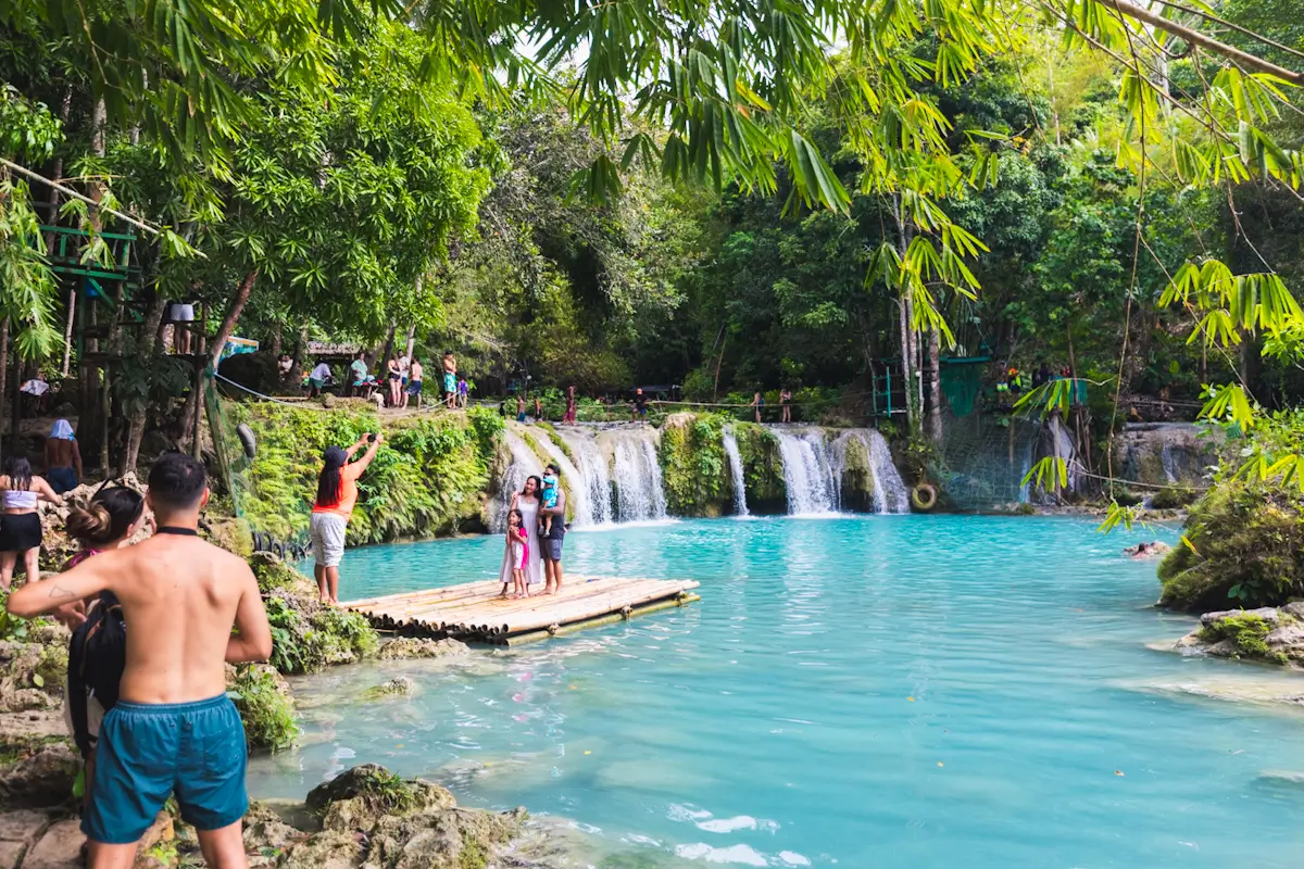 People stand on a wooden raft in a turquoise pool near the stunning Cambugahay Falls, a popular tourist attraction in Siquijor. Surrounded by lush green trees and rocks, visitors enjoy the serene natural scenery by the water and onshore.