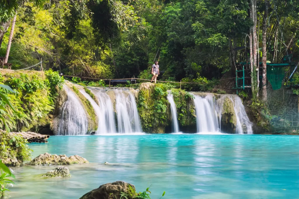 A person stands atop the clear blue waters of Cambugahay Falls, a popular tourist attraction in Siquijor, surrounded by lush green trees and vibrant foliage under daylight.